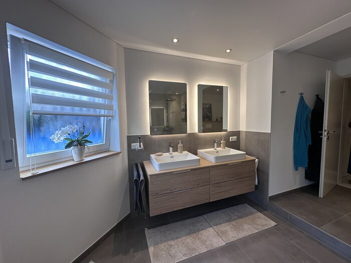 Badezimmer mit Doppelwaschtisch in Holzoptik und beleuchteten Spiegeln